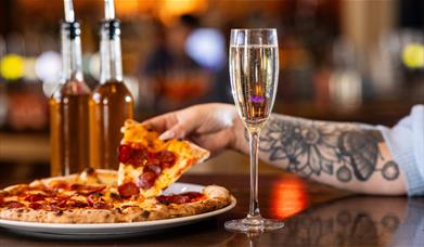 A hand lifting a slice of pizza from a plate with a glass of Prosecco in the foreground