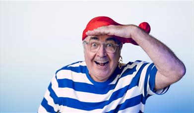 Danny Baker in a blue and white striped t-shirt and a red hat. He is holding his hand to his forehead.