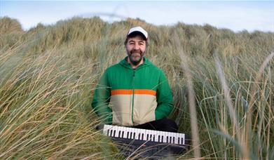 David O'Doherty sitting in tall grass with a small keyboard. Text reads "David O'Doherty, Tiny Piano Man"