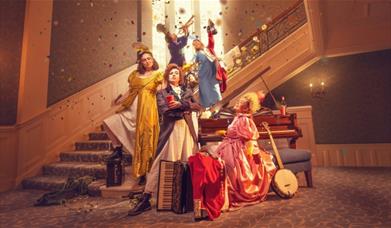 A group of women dressed in period costume. Three are at the bottom of a staircase grouped around a piano, while the other two are on the staircase, w