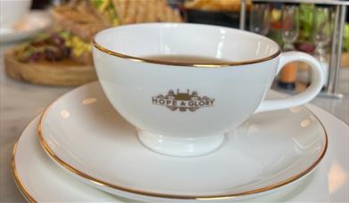 A white china with gold rim Hope & Glory branded tea cup and saucer with food in background at The Abbey Tea Bar, Bath