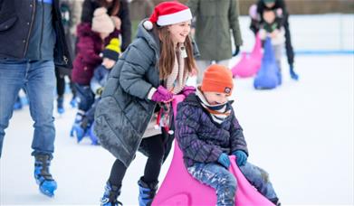 A young girl, wearing a Santa hat and winter coat, joyfully pushes a younger child on a pink ice skating aid while they both glide across the ice rink