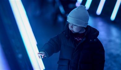 A young child, bundled up in a warm jacket and beanie, curiously touches a glowing light display. The scene captures a moment of wonder and fascinatio