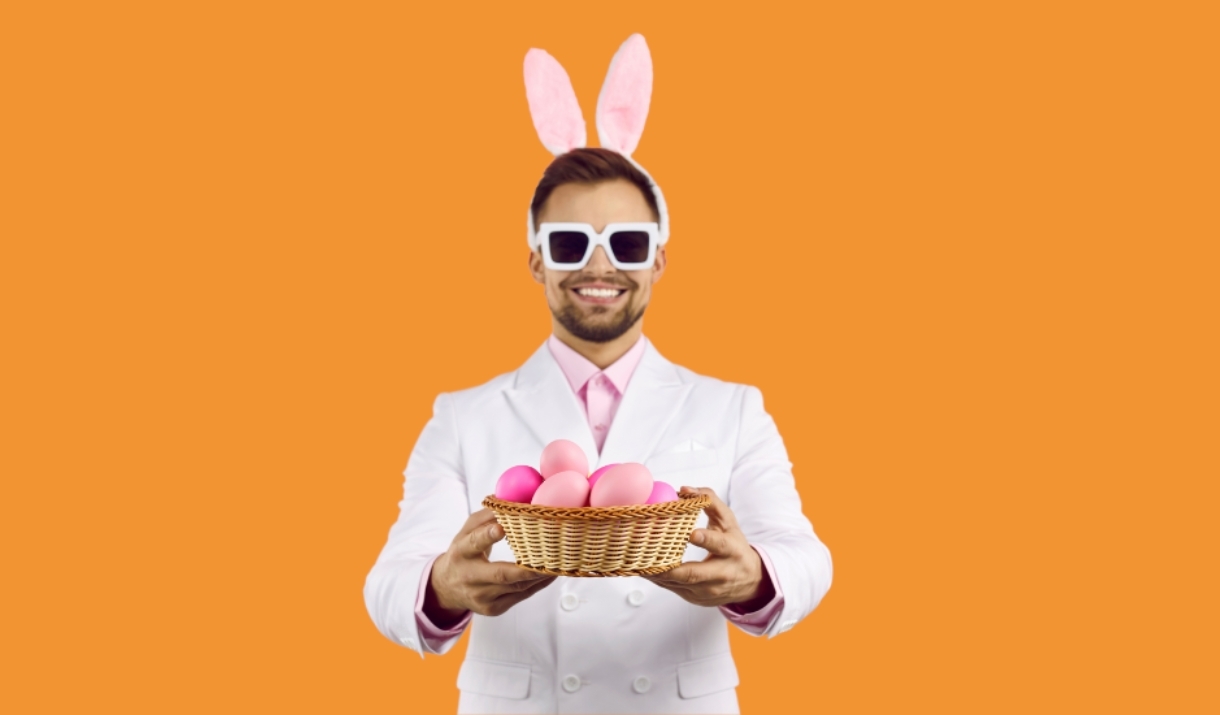 A cheerful man wearing a white suit, pink shirt, and bunny ears headband holds out a wicker basket filled with pink Easter eggs. He is also wearing ov