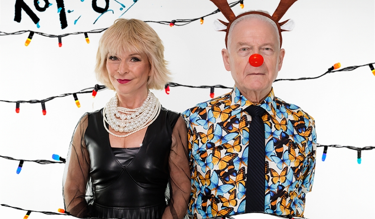 A photo of Toyah and Robert in front of a white background. Behind them are strings of Christmas lights. Toyah is wearing a black leather dress with a