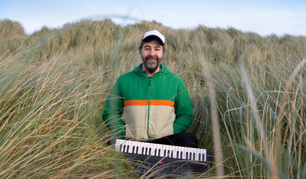 David O'Doherty sitting in tall grass with a small keyboard. Text reads "David O'Doherty, Tiny Piano Man"