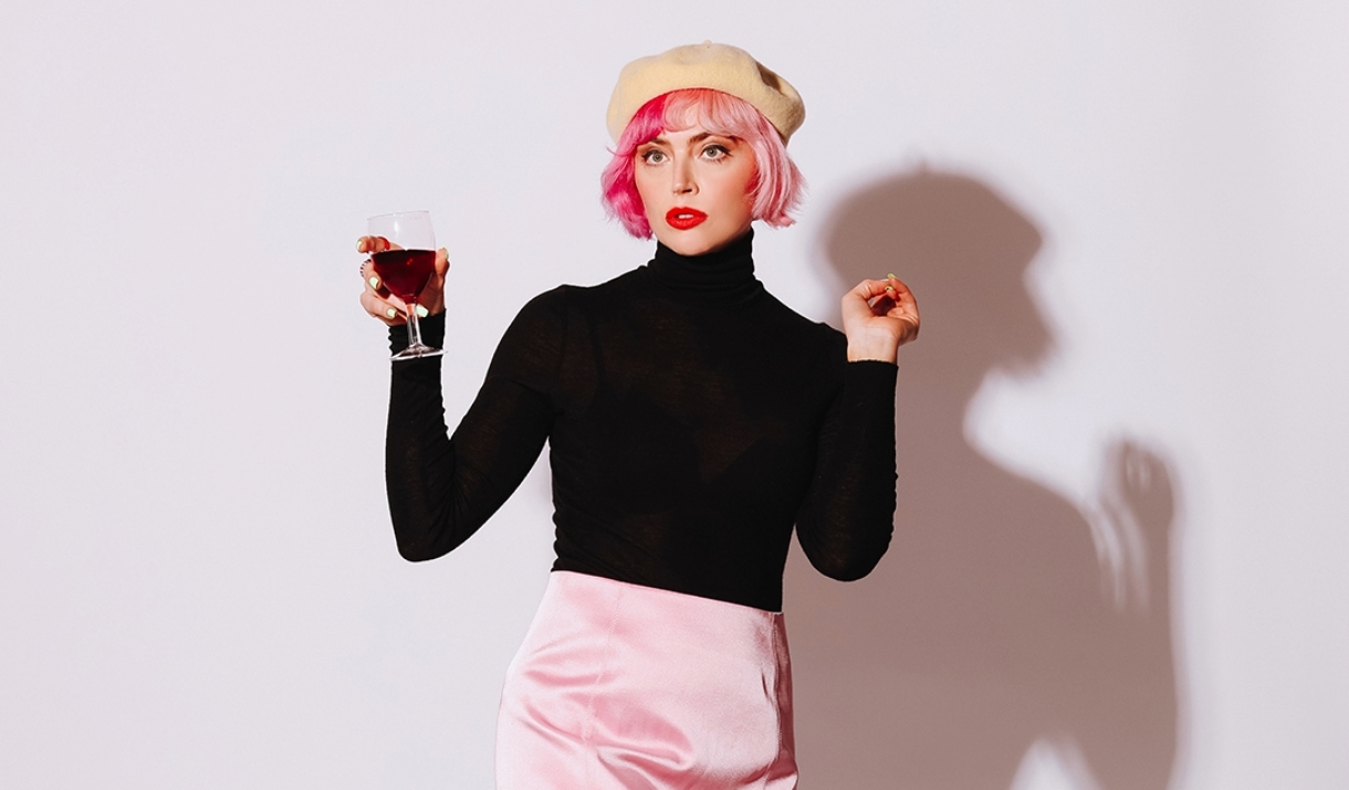 Photo of Tatty Macleod posing with a glass of red wine her hand. She has bright red lipstick and is wearing a light yellow beret, a black turtle neck