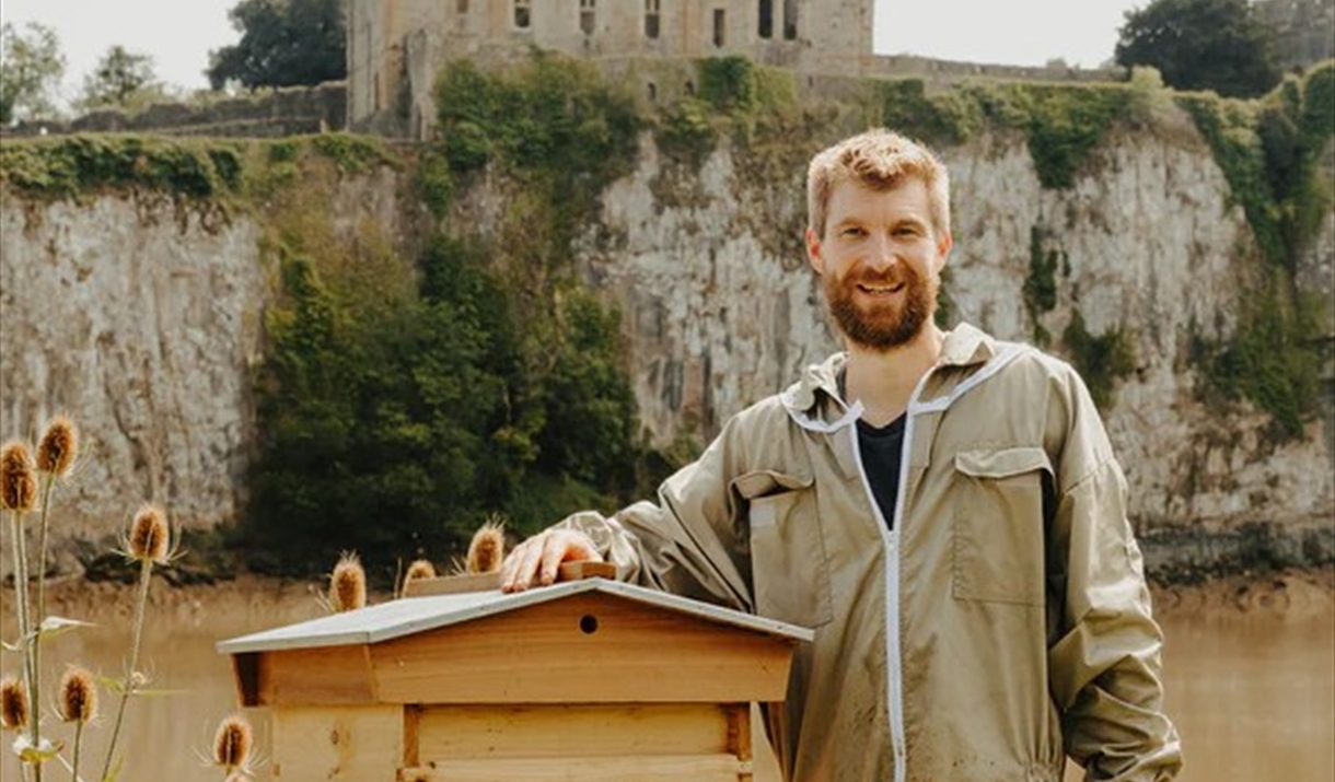 An Image of a bearded man in a beekeepers outfit standing beside a bee hive. There is a river behind him and a small abandoned church at the top of a