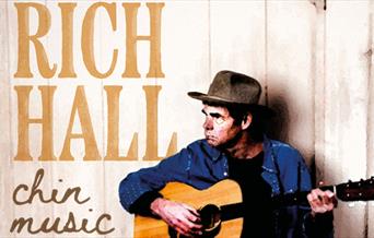 Rich Hall in a cowboy hat, playing an acoustic guitar