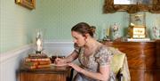 A woman in Regency clothing writing at a writing desk