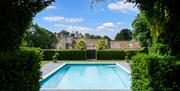 View of a pool through an archway made of hedges with lodging properties in the background. Blue sky with clouds.