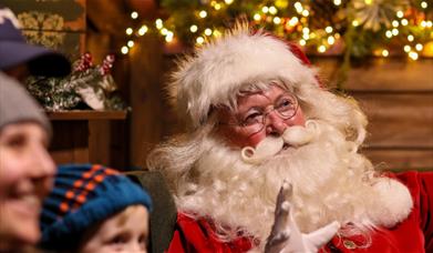 Santa Claus waving warmly at visitors, surrounded by festive holiday lights and decorations. The joyful scene captures the magic of Christmas, with Sa